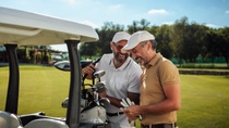 Two male friends laughing and talking by the golf cart on the field while getting ready to leave the golf court after a game