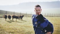 Farmer holding lamb in field with grazing sheep, illustrating sustainable farming through agroforestry integration. 