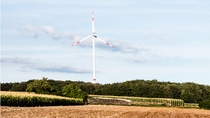 Wind turbine in rural landscape with fields and trees under blue sky. 