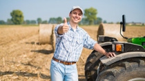 Smiling farmer standing by a tractor in a harvested field, giving a thumbs-up gesture. 