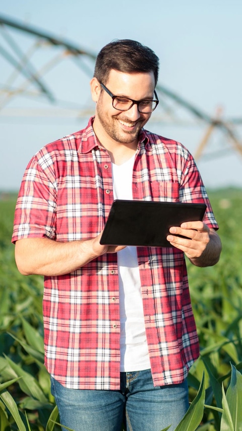 AR Feliz joven granjero usando tableta en el campo de maíz. Sistema de riego al fondo.